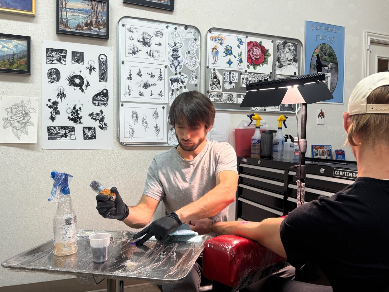 Cade Bennett tattooing at his station AK Tattoo Anchorage — flash sheets Alaska paintings